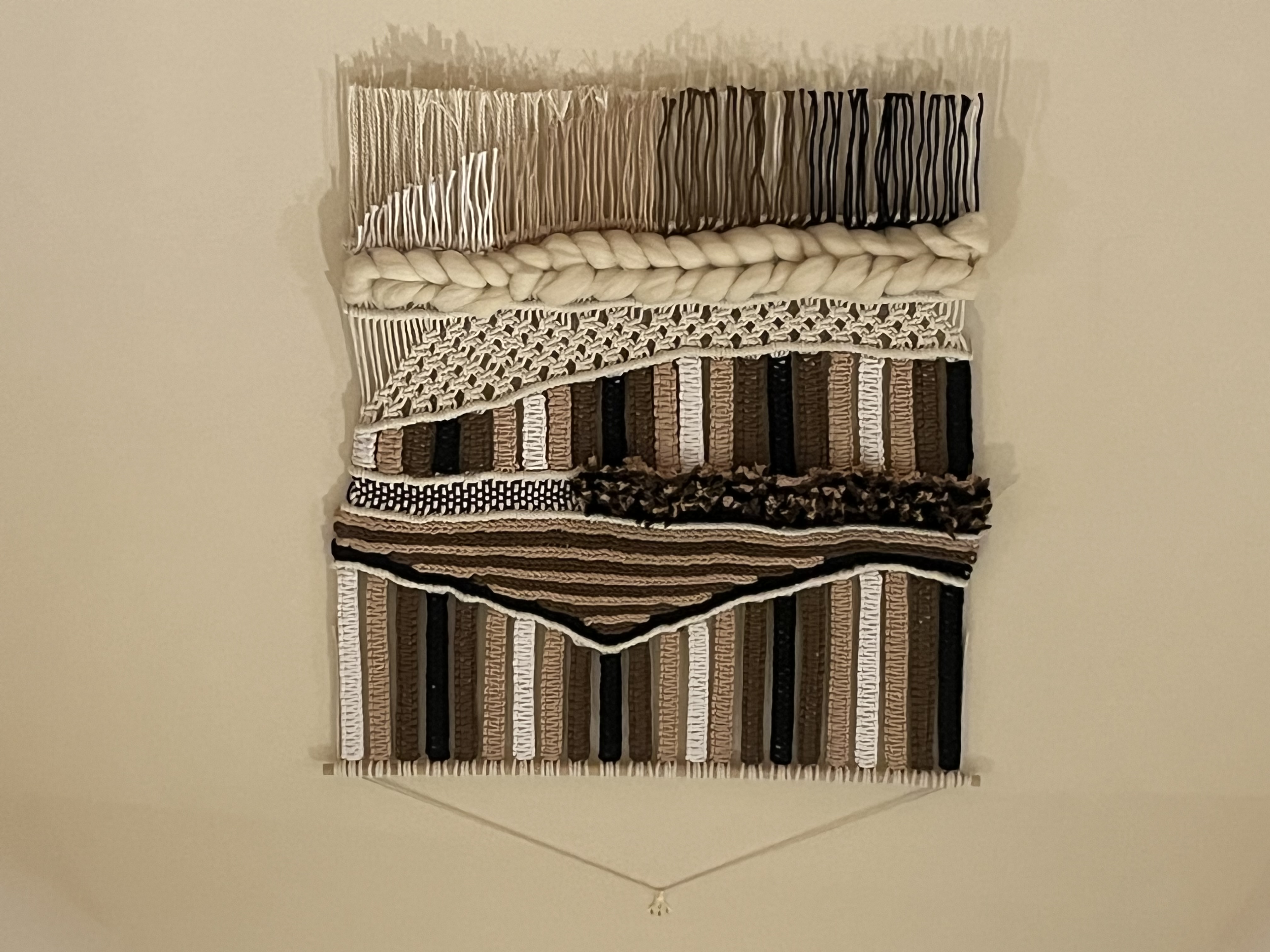 A handmade macramé wall hanging by Knot Perfect, featuring layered cream, tan and black fringing